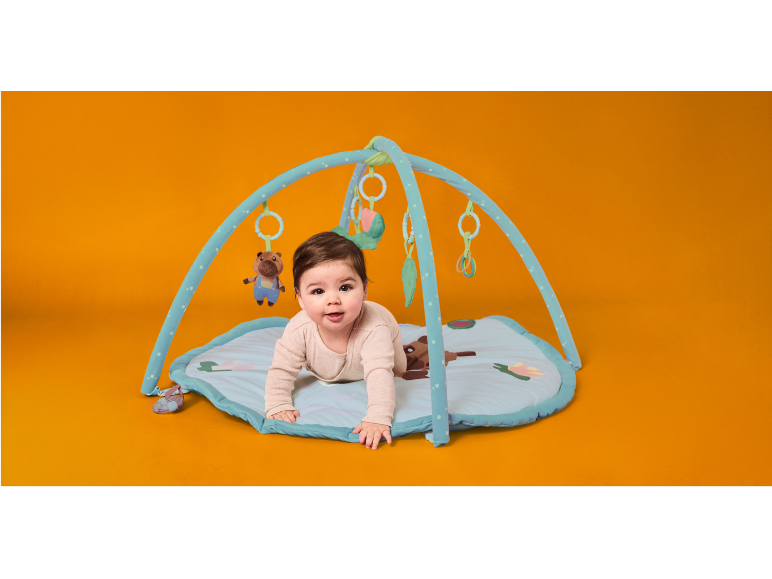 Bambino che gattona su un tappeto gioco con archi e giocattoli appesi su sfondo arancione.