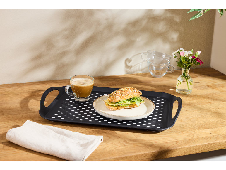 Vassoio nero con caffè e panino su un tavolo di legno, con fiori e tazze.