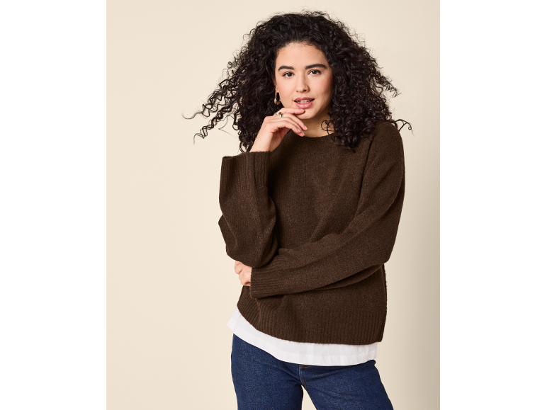 Donna con maglione marrone e jeans, con la mano sul mento.