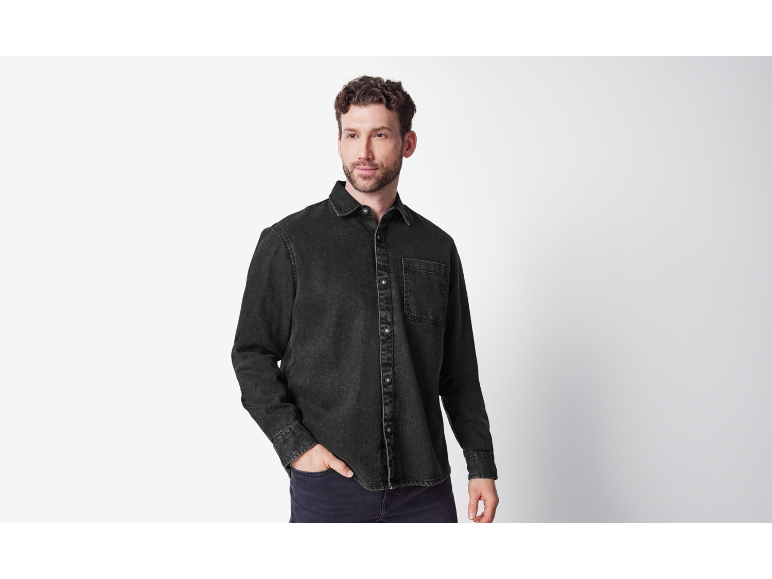 Uomo con camicia di jeans nera e pantaloni scuri, in posa con la mano in tasca.