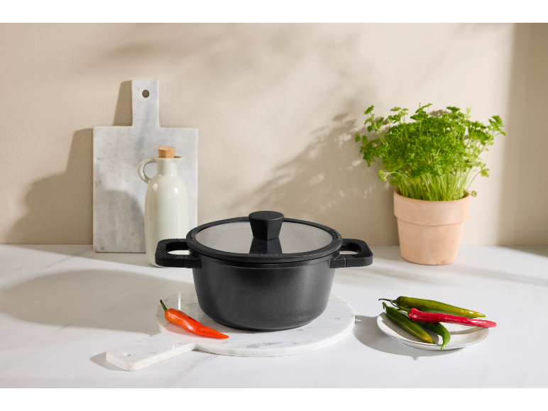 Pentola nera con coperchio in vetro, peperoncini ed erbe fresche su un bancone da cucina.