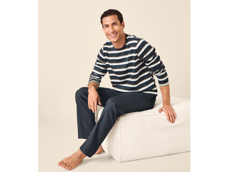 Uomo sorridente in pigiama a righe e pantaloni blu scuro, seduto su un pouf bianco.