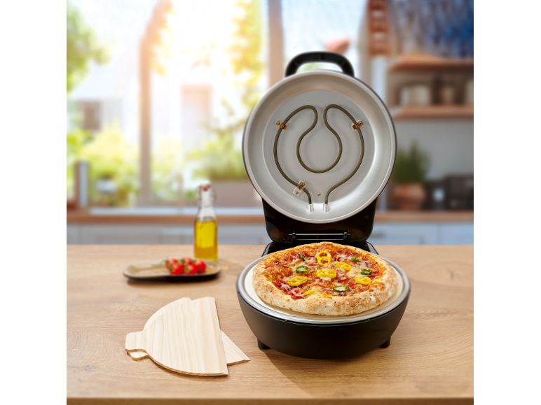 Forno elettrico per pizza aperto con pizza appena fatta e palette di legno in una cucina luminosa.