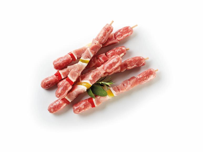 Spiedini di salsiccia e pancetta con peperoni e salvia su sfondo bianco.