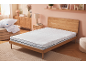 Camera da letto con materasso bianco, cuscini e piumino su un pouf, e mobili in legno.