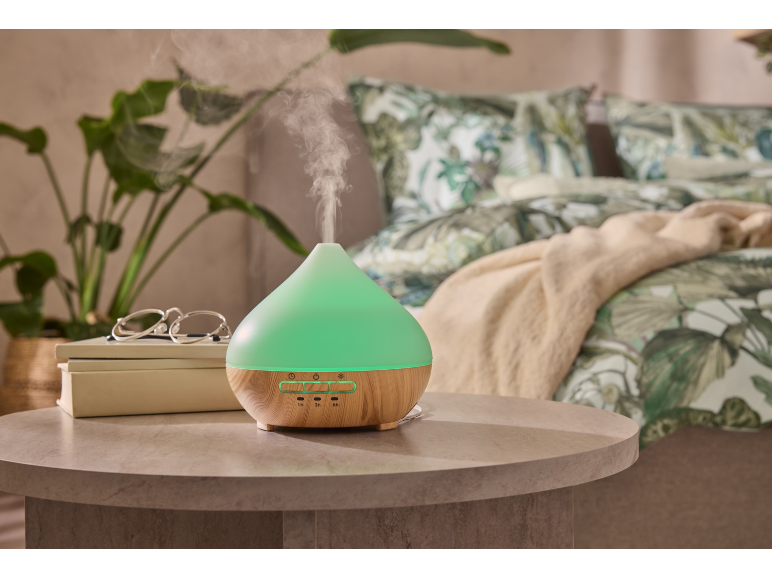 Diffusore di aromi verde con base in legno in una camera da letto con piante e biancheria da letto a fantasia.