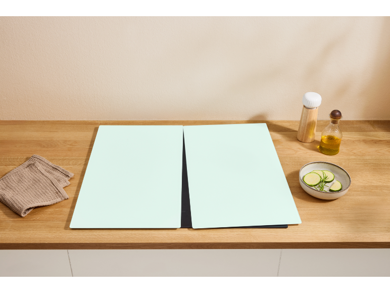 Piano di lavoro in legno con due taglieri verde chiaro, un panno, olio e una ciotola di zucchine.