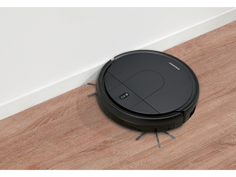Robot aspirapolvere nero che pulisce un pavimento in legno accanto a una parete bianca.