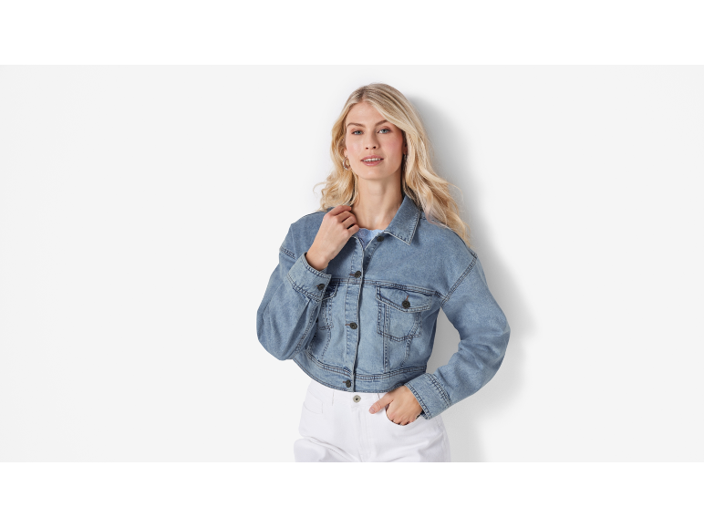 Donna in giacca di jeans blu e pantaloni bianchi, in posa con la mano in tasca.