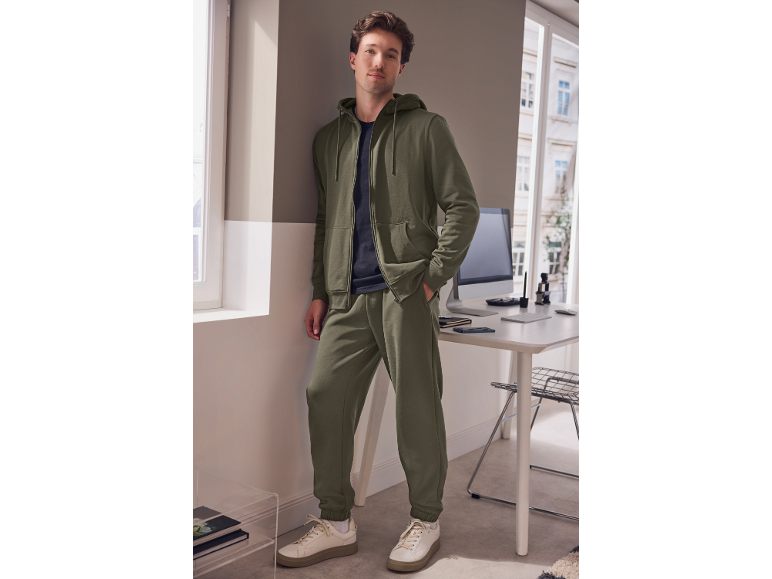 Uomo in tuta verde oliva e sneakers, appoggiato a una parete.