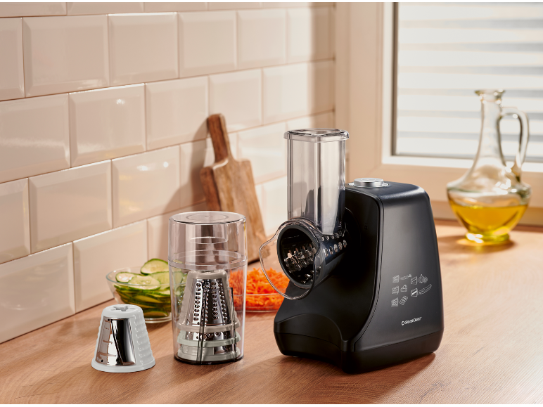 Robot da cucina nero con accessori per affettare e grattugiare, con verdure preparate.
