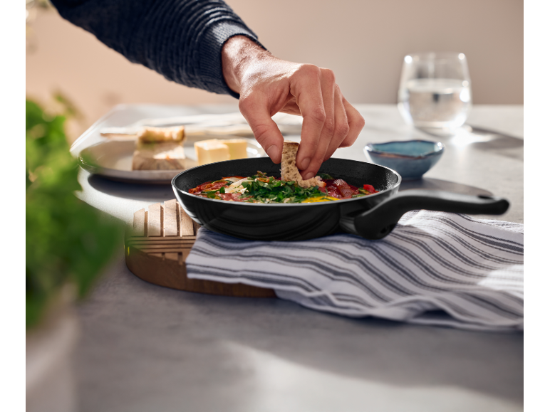 Mano di uomo che intinge il pane in una padella di shakshuka, con formaggio e pane a lato.