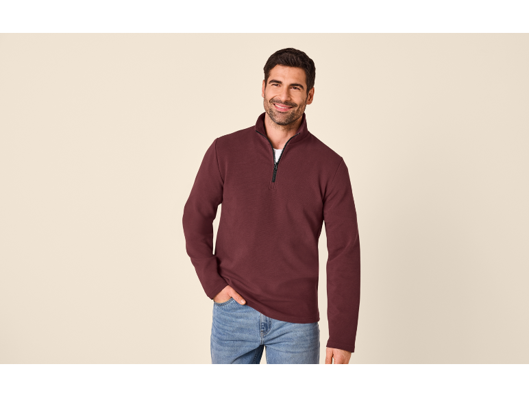 Uomo con felpa bordeaux a mezza zip e jeans, sorridente.