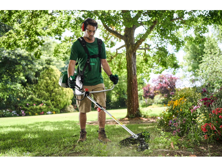 Uomo con imbracatura e cuffie antirumore che taglia l'erba con un decespugliatore in giardino.