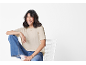 Donna sorridente con maglietta beige e jeans blu, seduta su una sedia bianca.