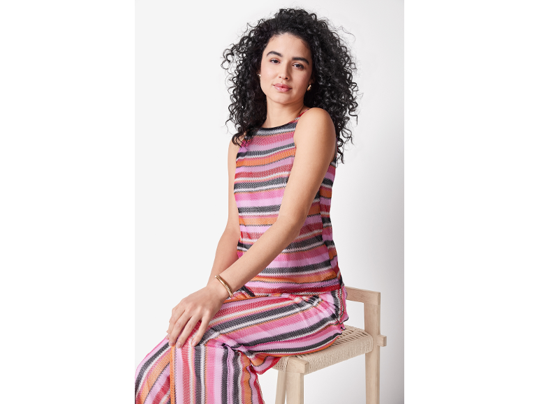 Donna con capelli ricci, top e pantaloni a righe colorate, seduta su uno sgabello.