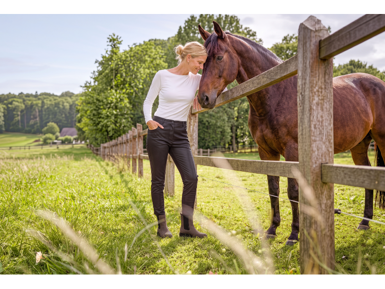 Donna in pantaloni neri e top bianco accarezza un cavallo marrone vicino a una recinzione di legno.