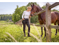 Donna in pantaloni neri e top bianco accarezza un cavallo marrone vicino a una recinzione di legno.