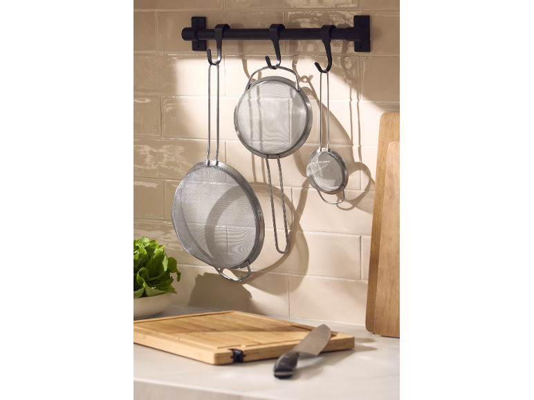 Colini da cucina in acciaio inossidabile appesi a una barra a muro sopra un tagliere.