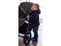 Bambino in giacca a vento blu navy e jeans, in piedi accanto a un passeggino grigio scuro.