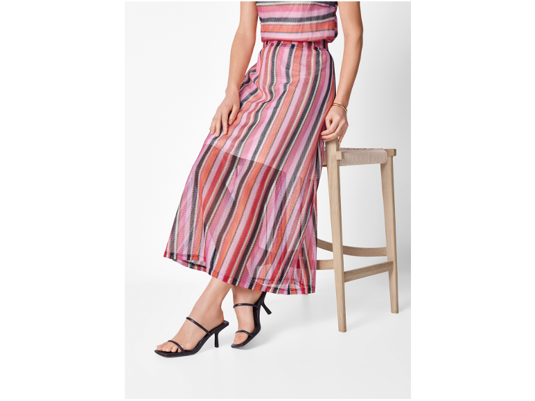 Donna in abito a righe colorate e sandali neri con tacco, appoggiata a uno sgabello.