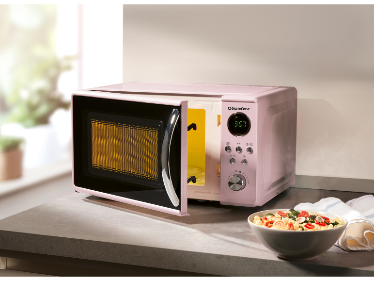 Forno a microonde rosa con porta aperta e ciotola di pasta sul bancone della cucina.