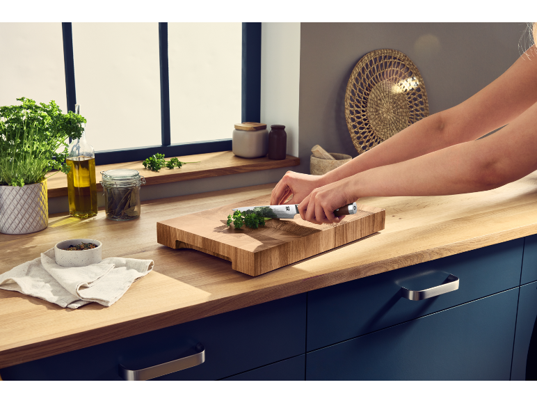 Persona che trita prezzemolo su un tagliere di legno in cucina con erbe e olio.