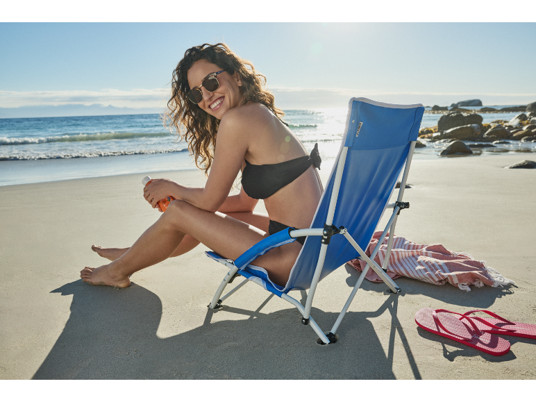 Donna sorridente in costume da bagno seduta su una sedia da spiaggia con crema solare.