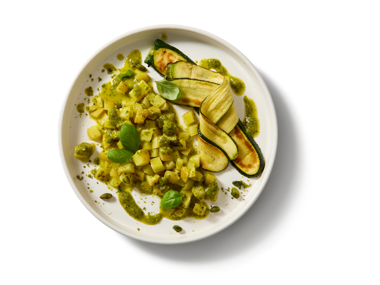 Patate e zucchine grigliate con salsa pesto e foglie di basilico su un piatto bianco.