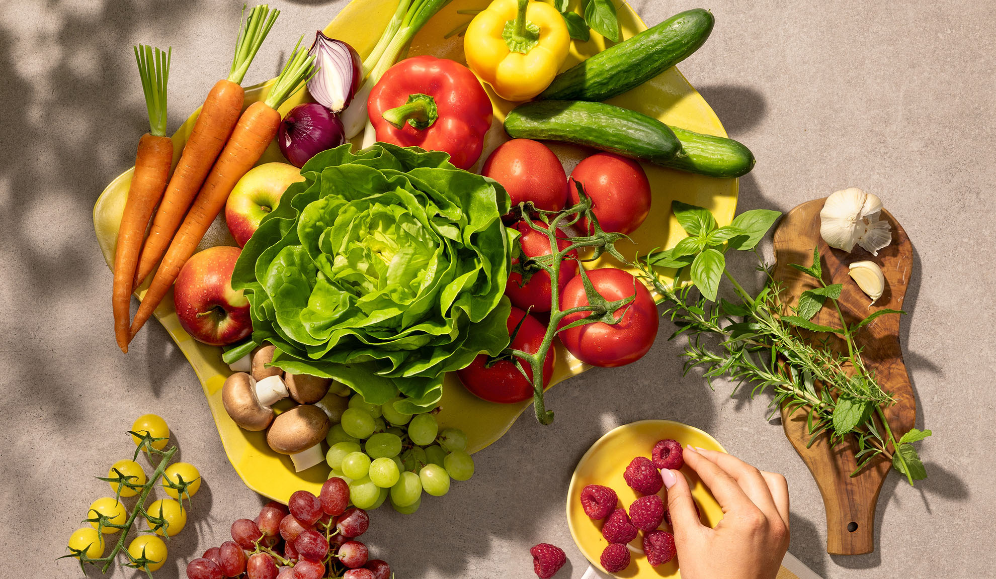 Verdure e frutta fresca su un piatto giallo, tra cui carote, lattuga, peperoni, pomodori e uva.