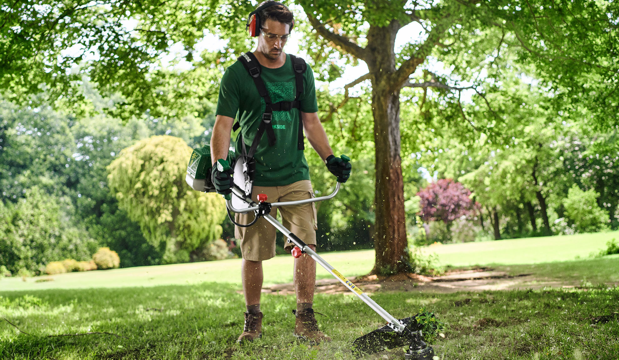 Uomo taglia l'erba in giardino con un decespugliatore, indossa occhiali e cuffie protettive.