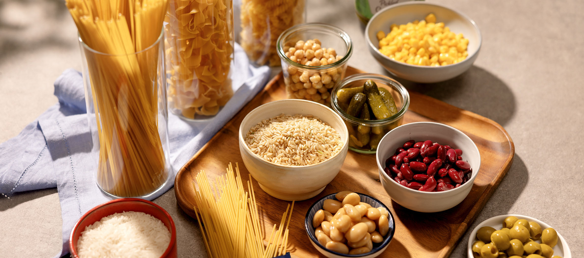 Vari prodotti base della dispensa, tra cui pasta, riso, fagioli, ceci, mais, sottaceti e olive.