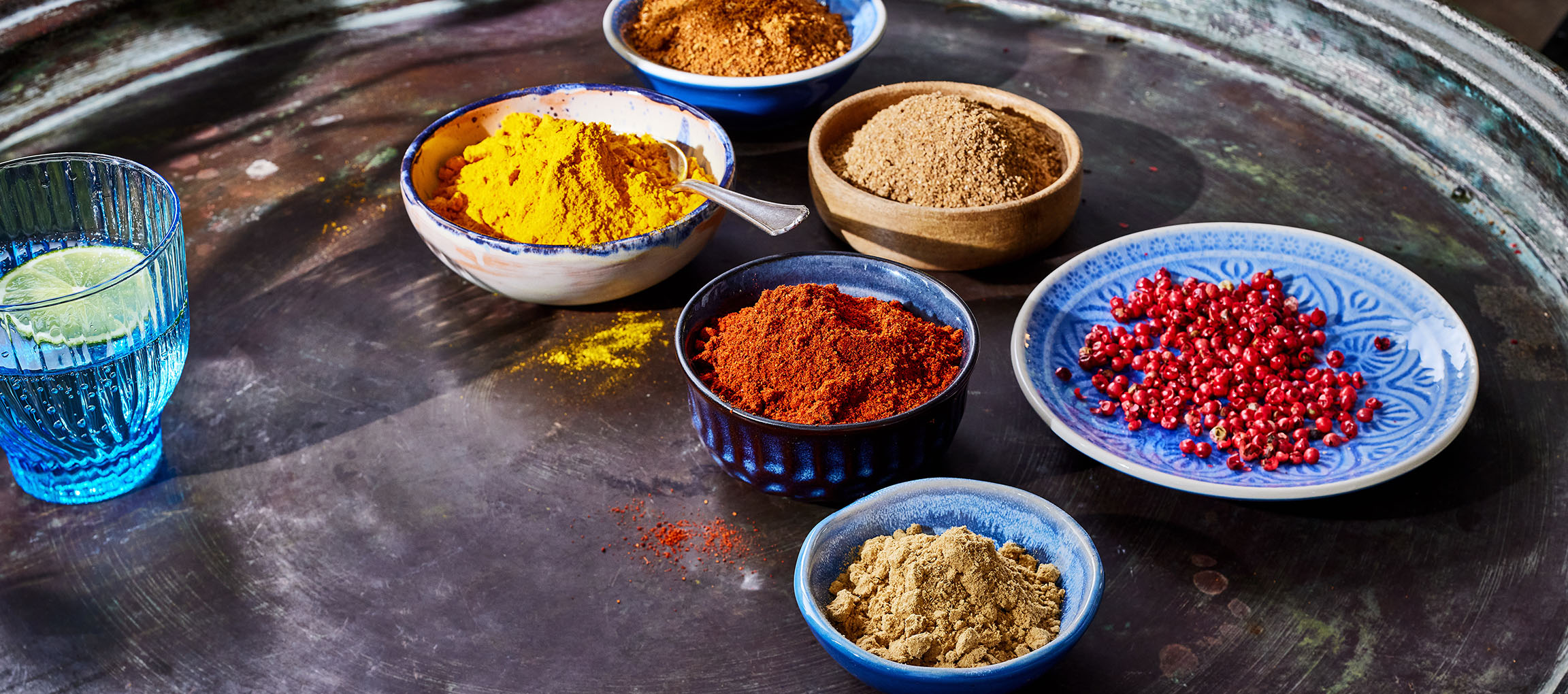 Varie spezie, tra cui curcuma, paprika e pepe rosa, in ciotole su una superficie scura.