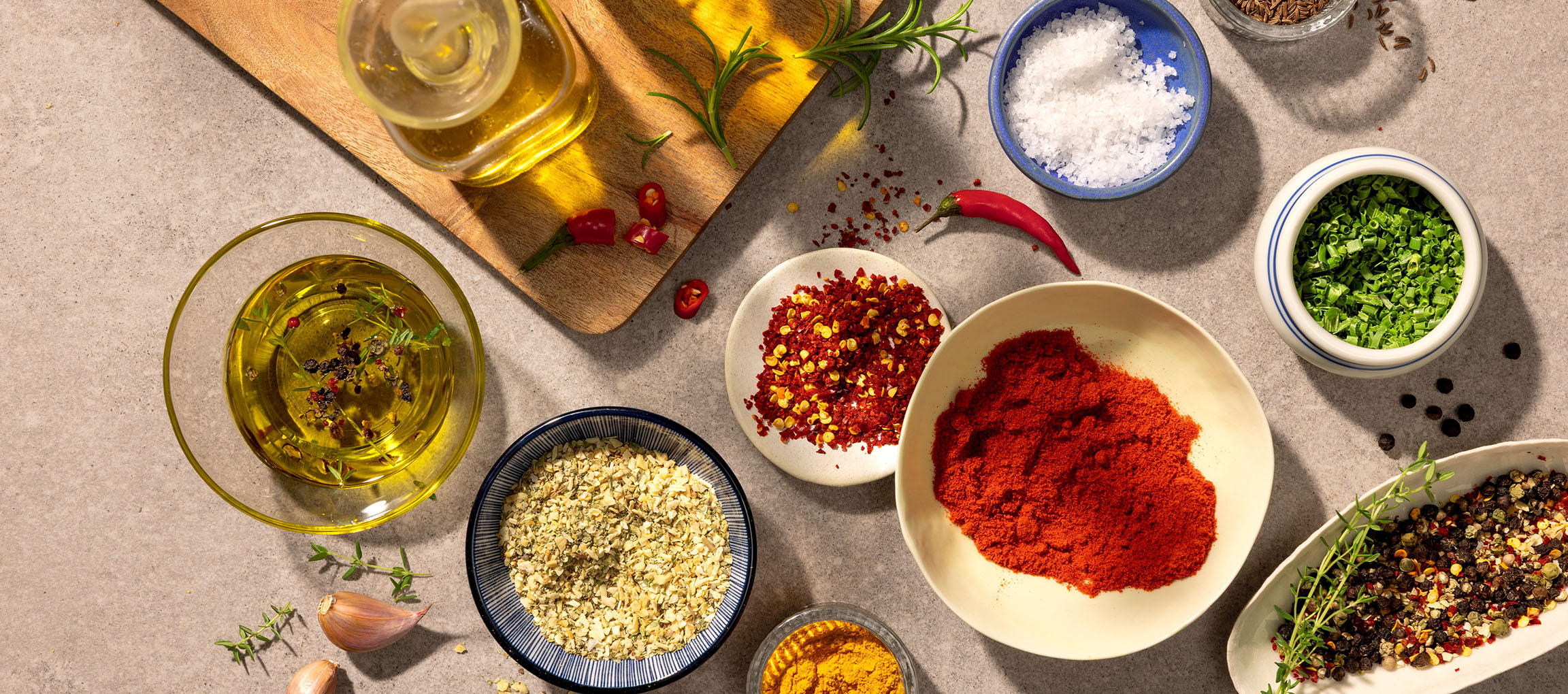 Spezie e oli vari, tra cui paprika, peperoncino, sale, pepe e olio d'oliva, su sfondo grigio.