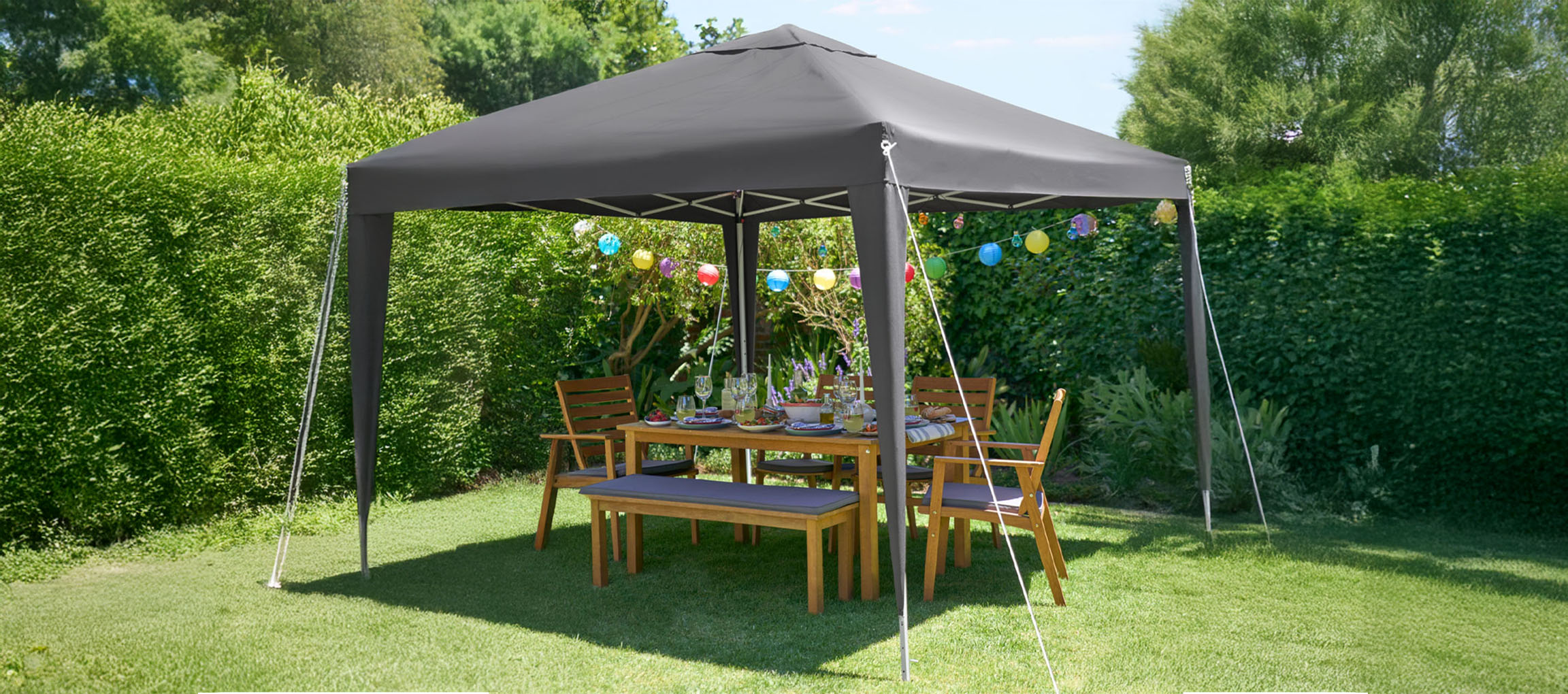 Un gazebo apribile grigio scuro protegge un set da pranzo in legno per esterni in un giardino verde lussureggiante.