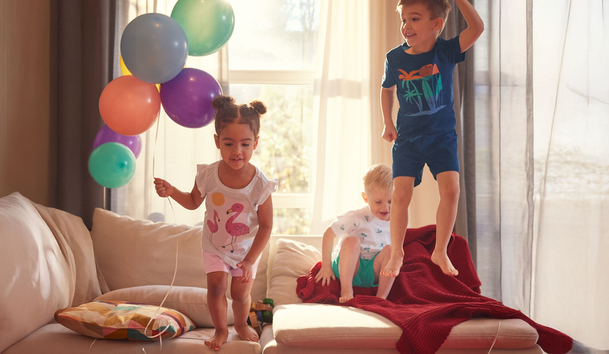 Tre bambini saltano allegramente su un divano con palloncini colorati.
