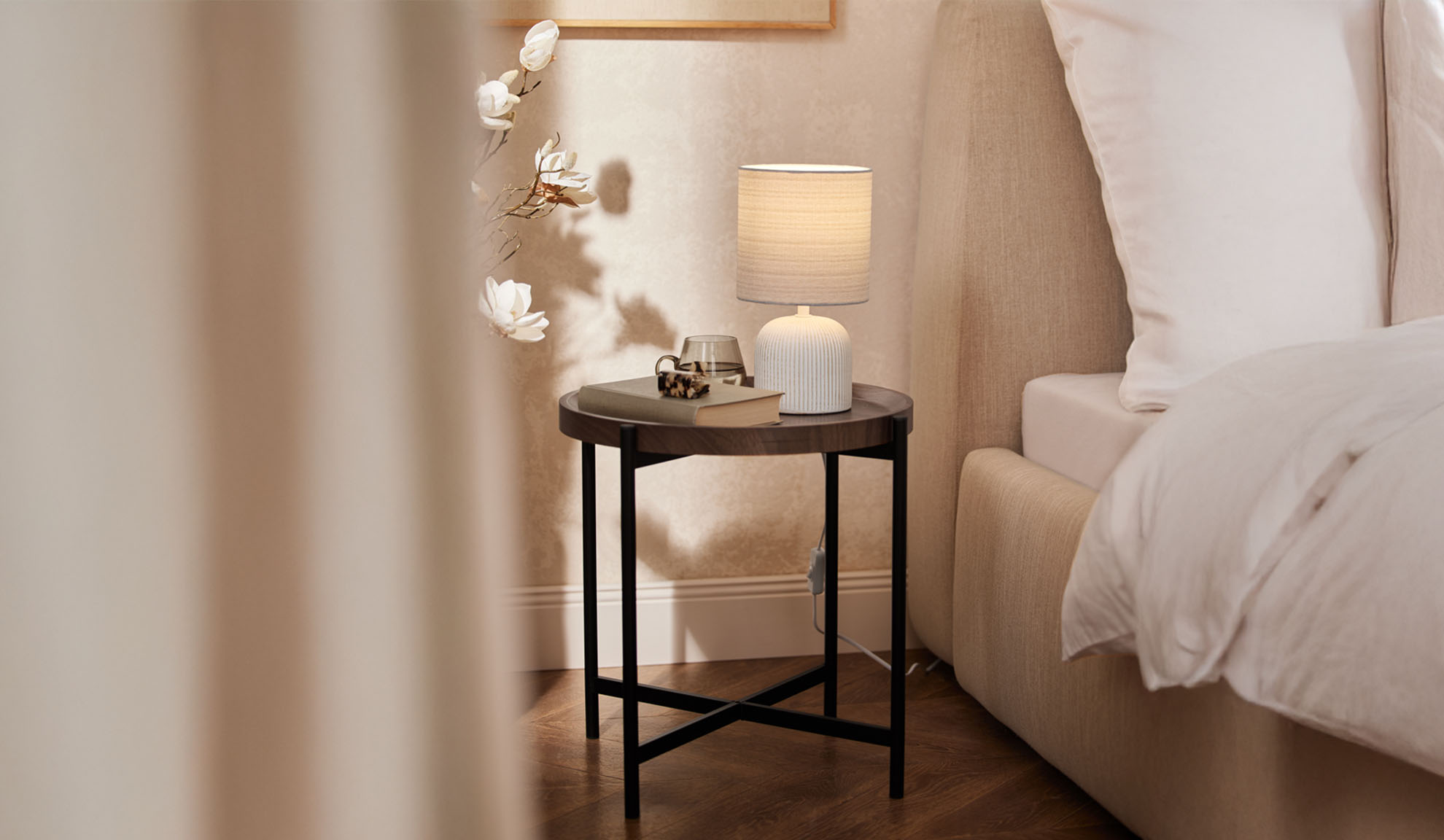 Comodino rotondo in legno con lampada da tavolo e accessori accanto a un letto.