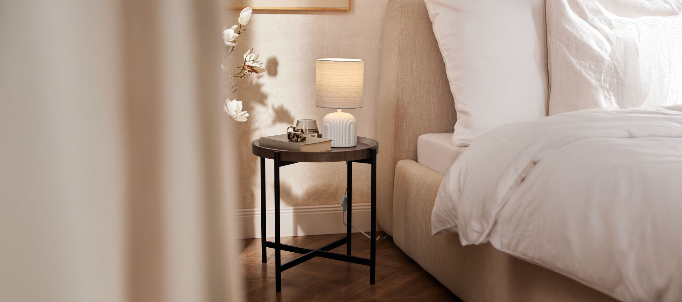 Comodino rotondo in legno con lampada da tavolo e letto beige in una camera da letto accogliente.