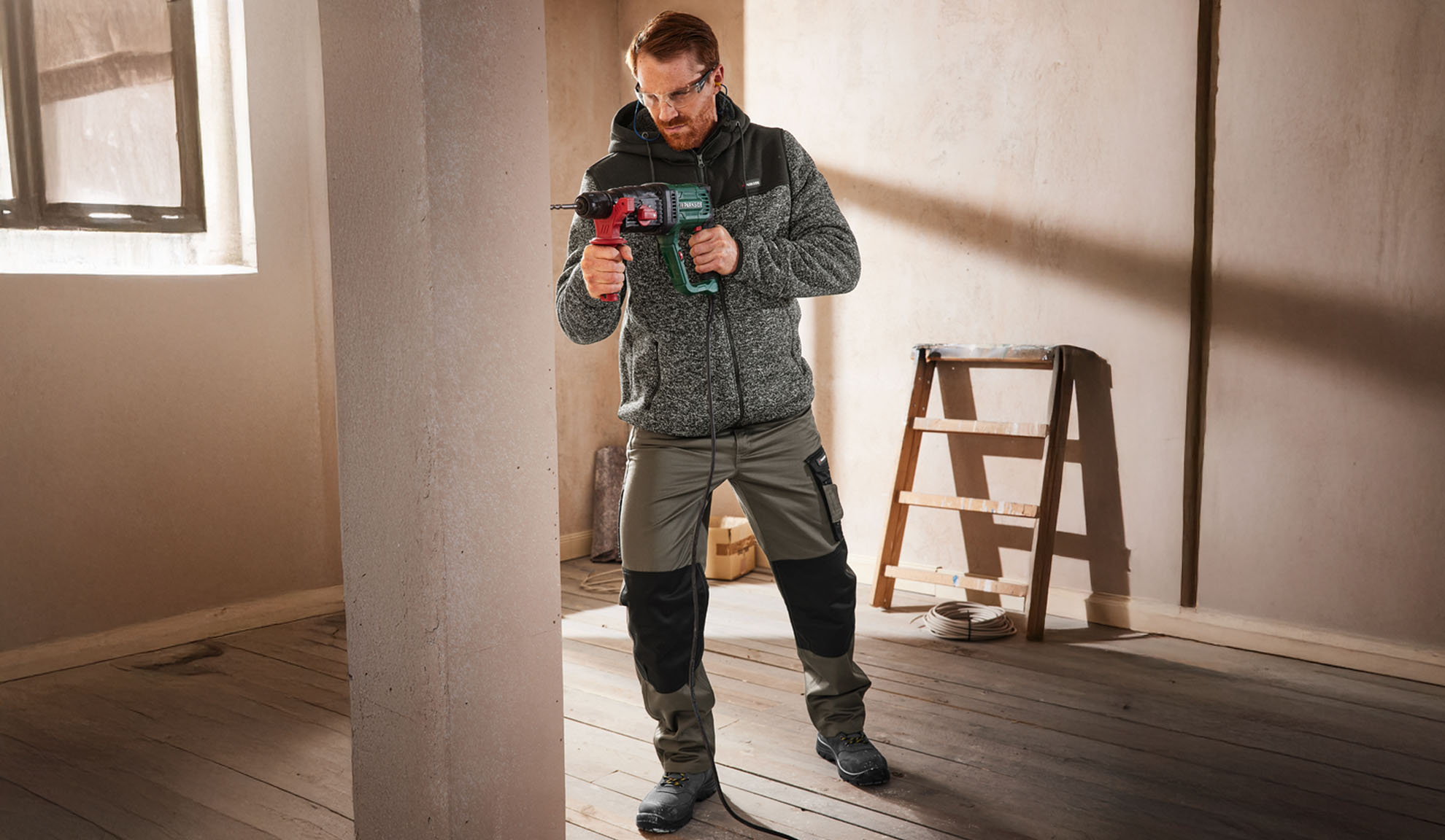 Uomo con giacca e pantaloni da lavoro, tiene un trapano in una stanza non finita.