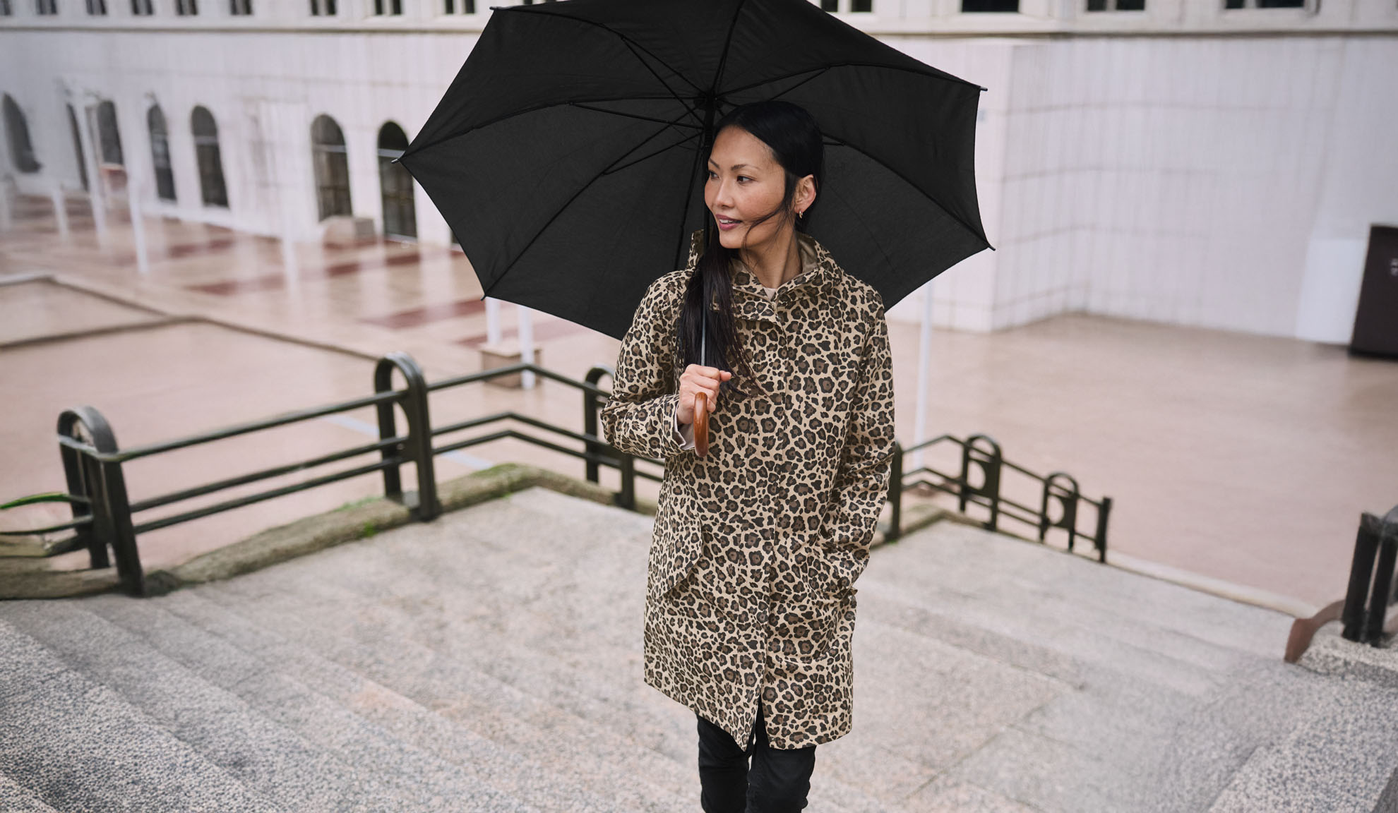 Una donna con un cappotto leopardato tiene un ombrello nero su una scalinata.