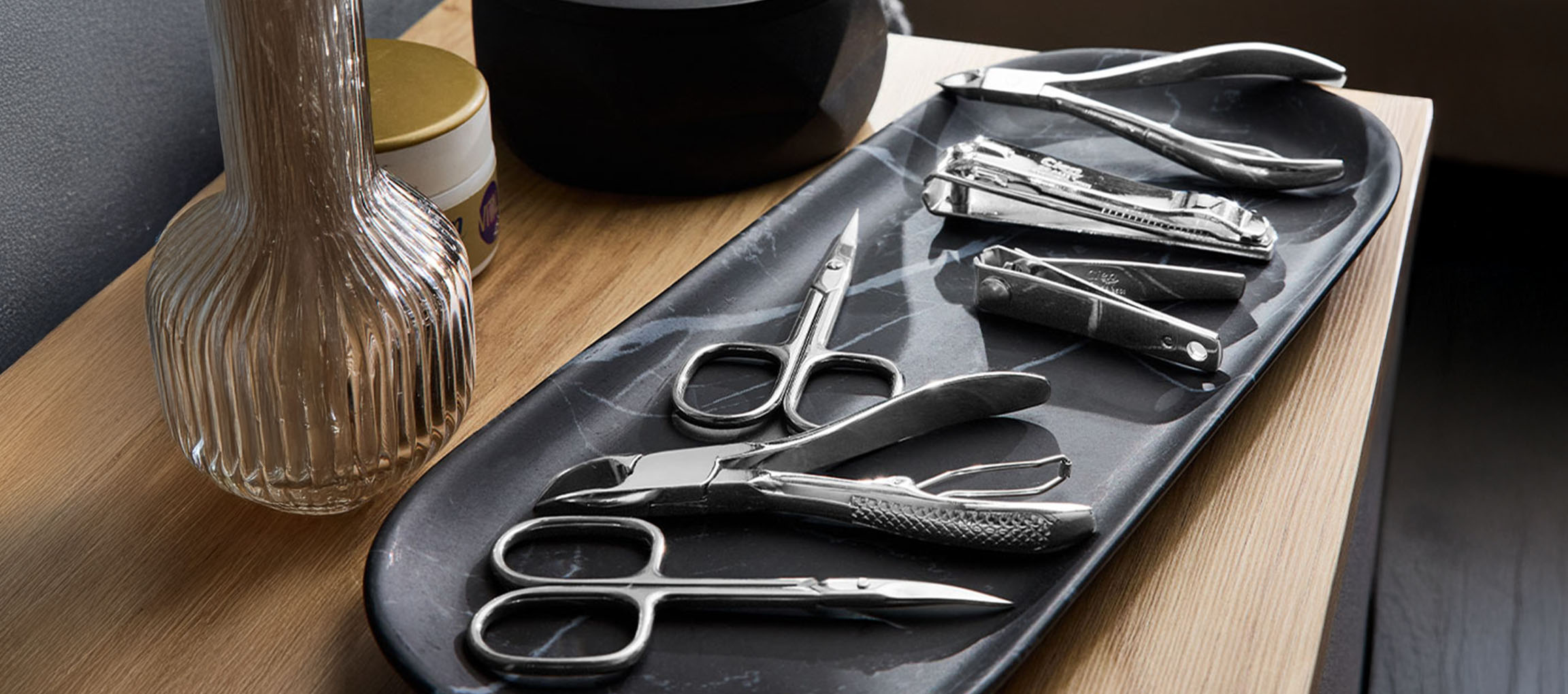 Strumenti per manicure e pedicure su un vassoio di marmo, con un vaso e un barattolo sullo sfondo.