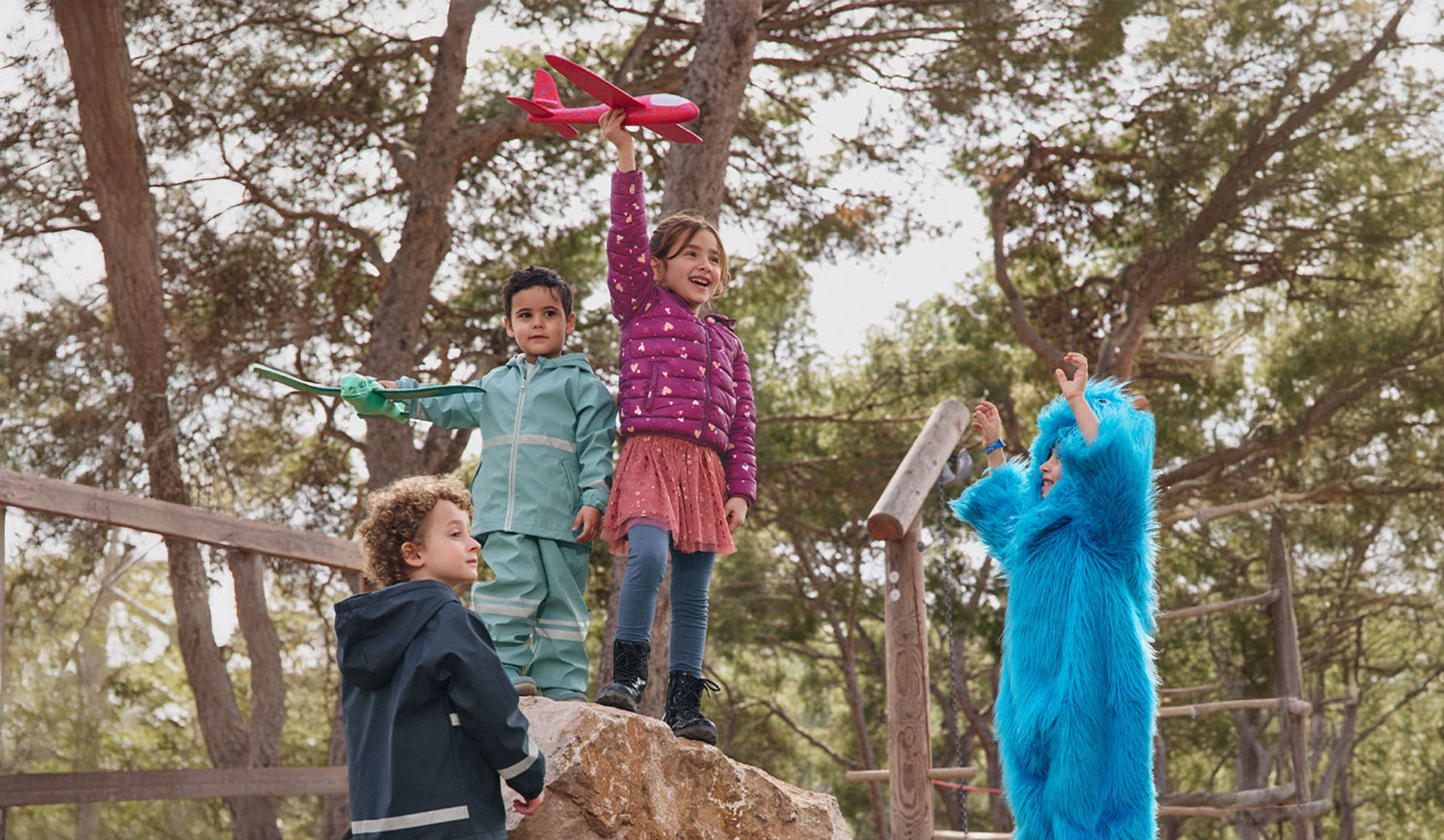 Quattro bambini giocano all'aperto, due con aeroplani giocattolo e uno in costume da mostro blu.