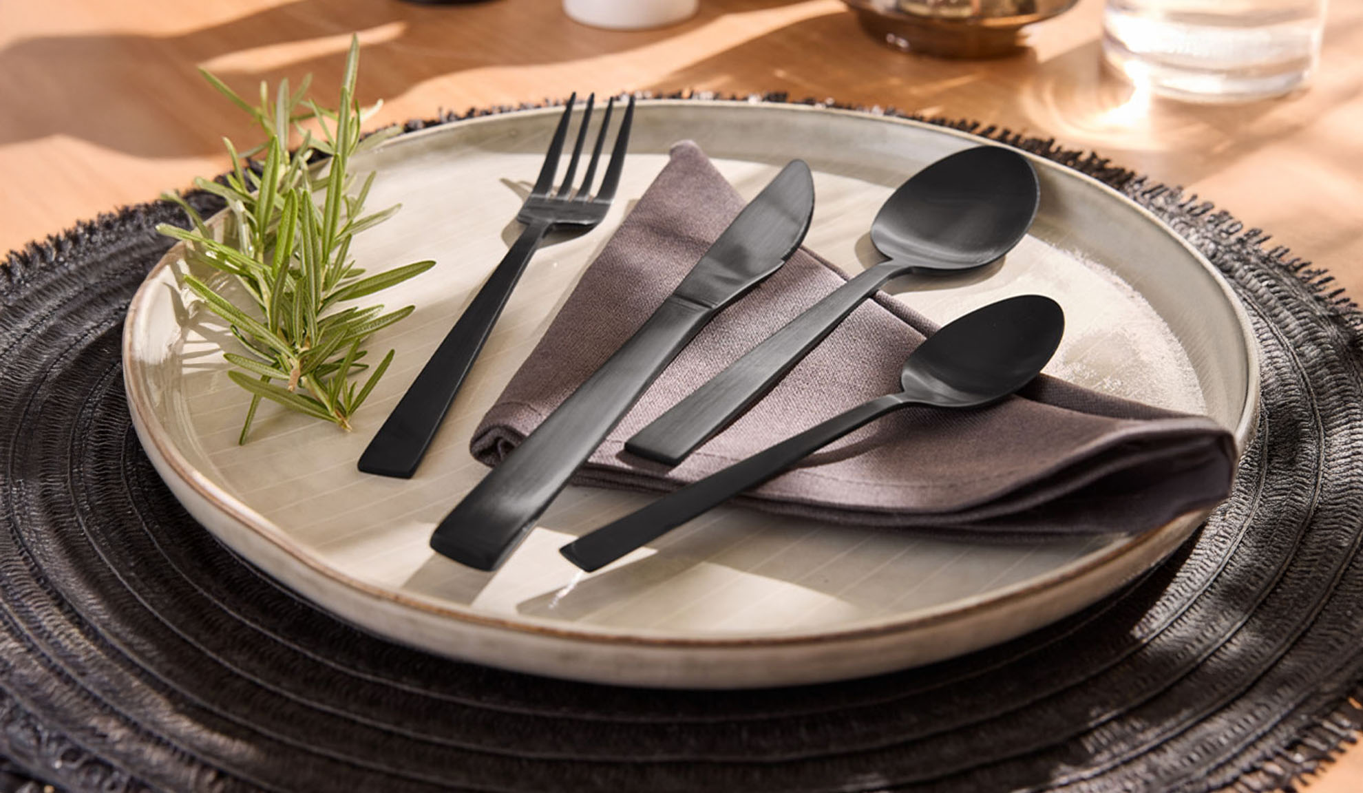 Set di posate nere con piatto, tovagliolo e rametto di rosmarino su tovaglietta.