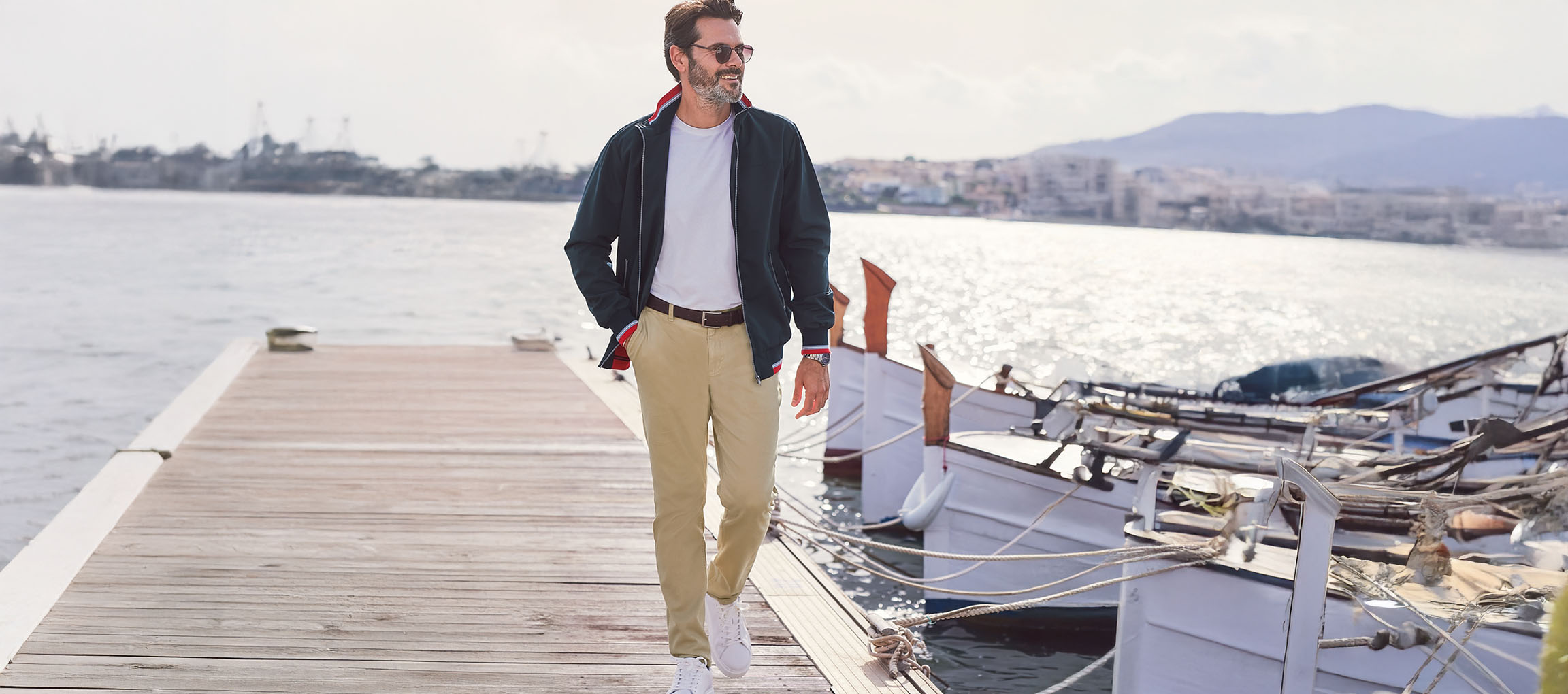 Uomo con giacca bomber verde scuro, pantaloni chino e sneakers bianche su un molo.