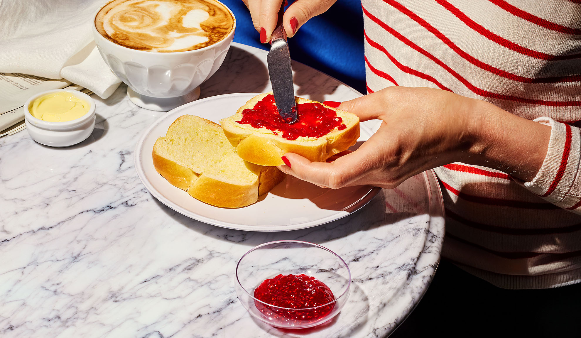 Mani che spalmano marmellata su pane brioche, con caffè e burro su tavolo di marmo.