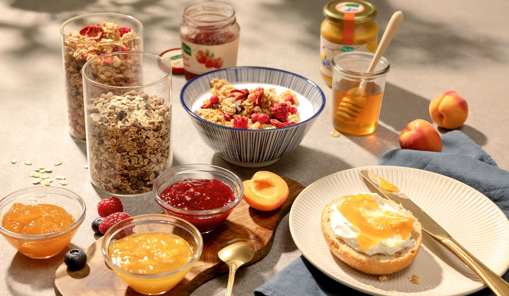 Colazione con granola, yogurt, miele, marmellate e frutta fresca su un tavolo soleggiato.