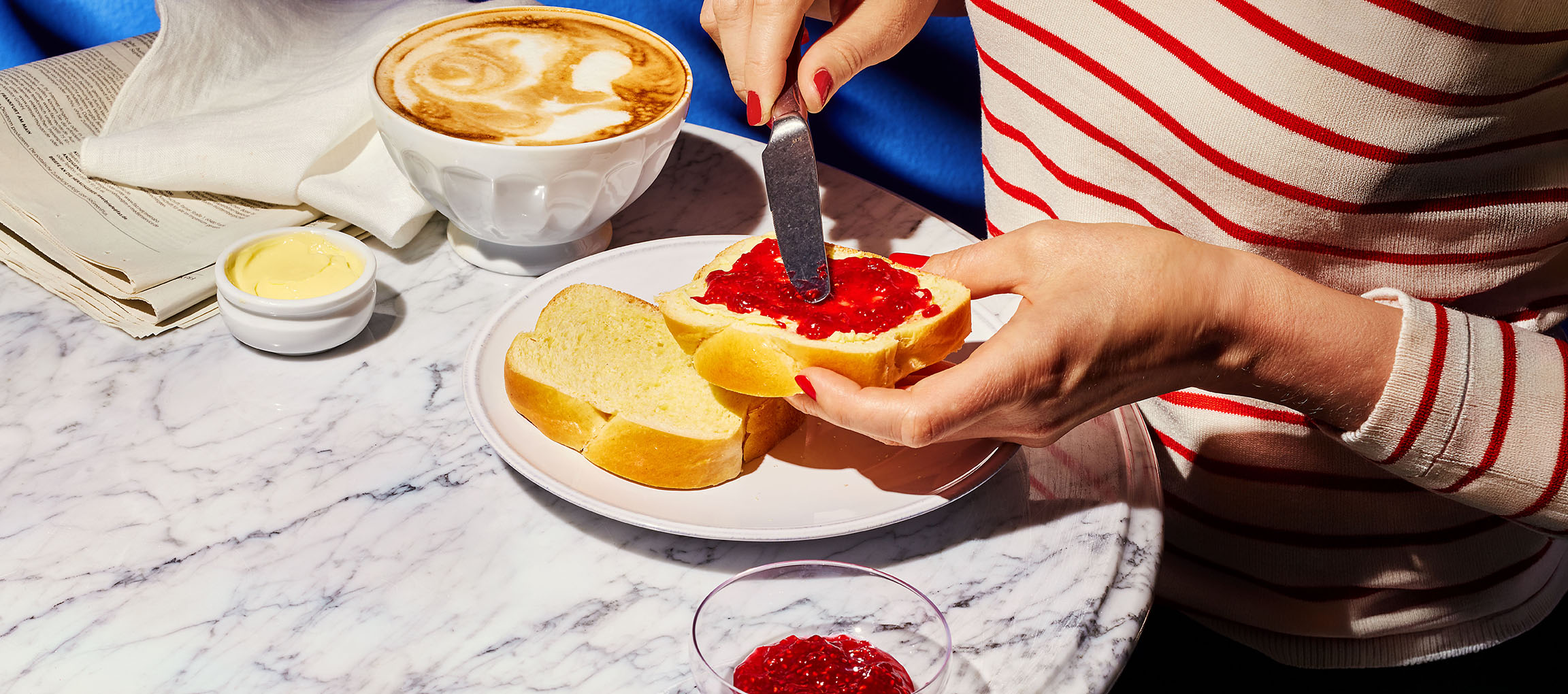 Mani che spalmano marmellata su pane brioche, con caffè e burro su tavolo di marmo.