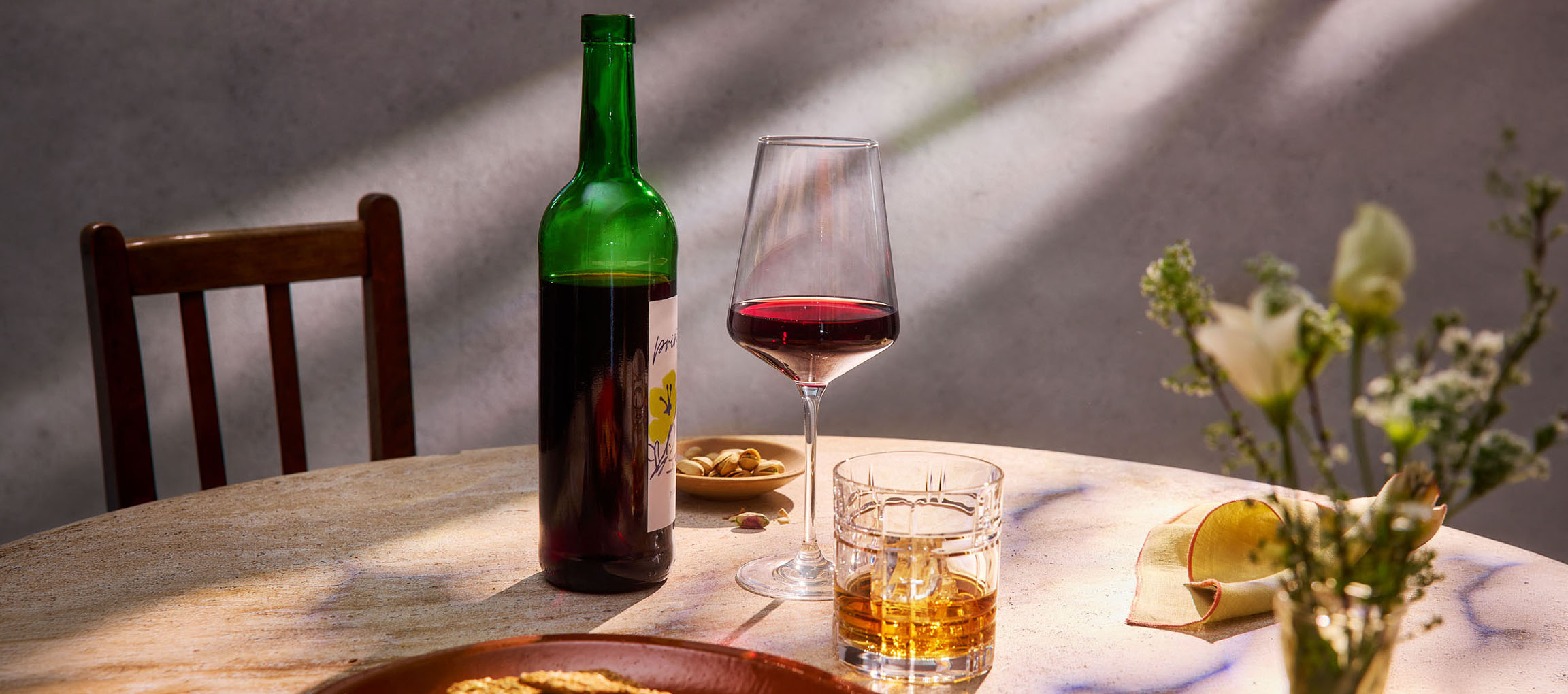 Bottiglia di vino rosso, bicchiere di vino e whisky su un tavolo, con sedia e fiori.