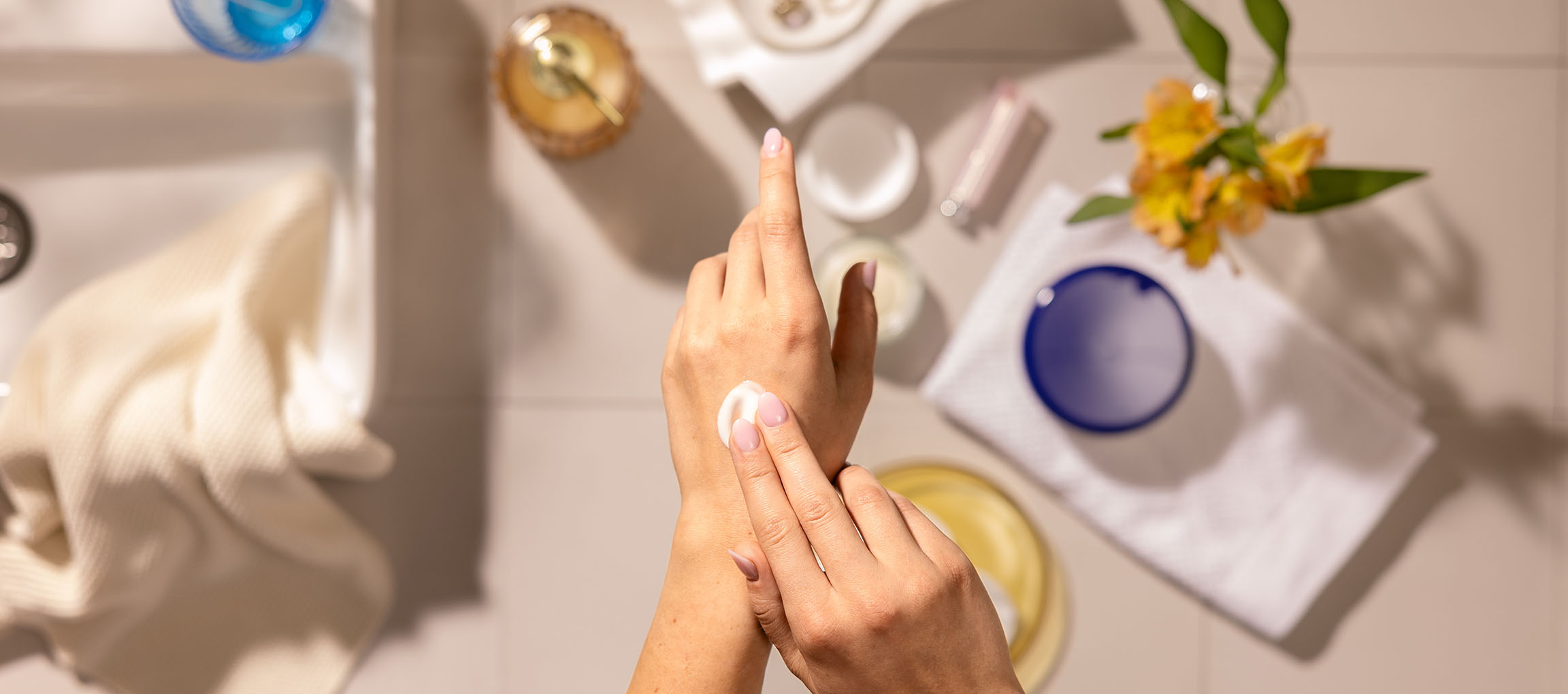 Mani femminili applicano crema per le mani, circondate da cosmetici e asciugamani in bagno.