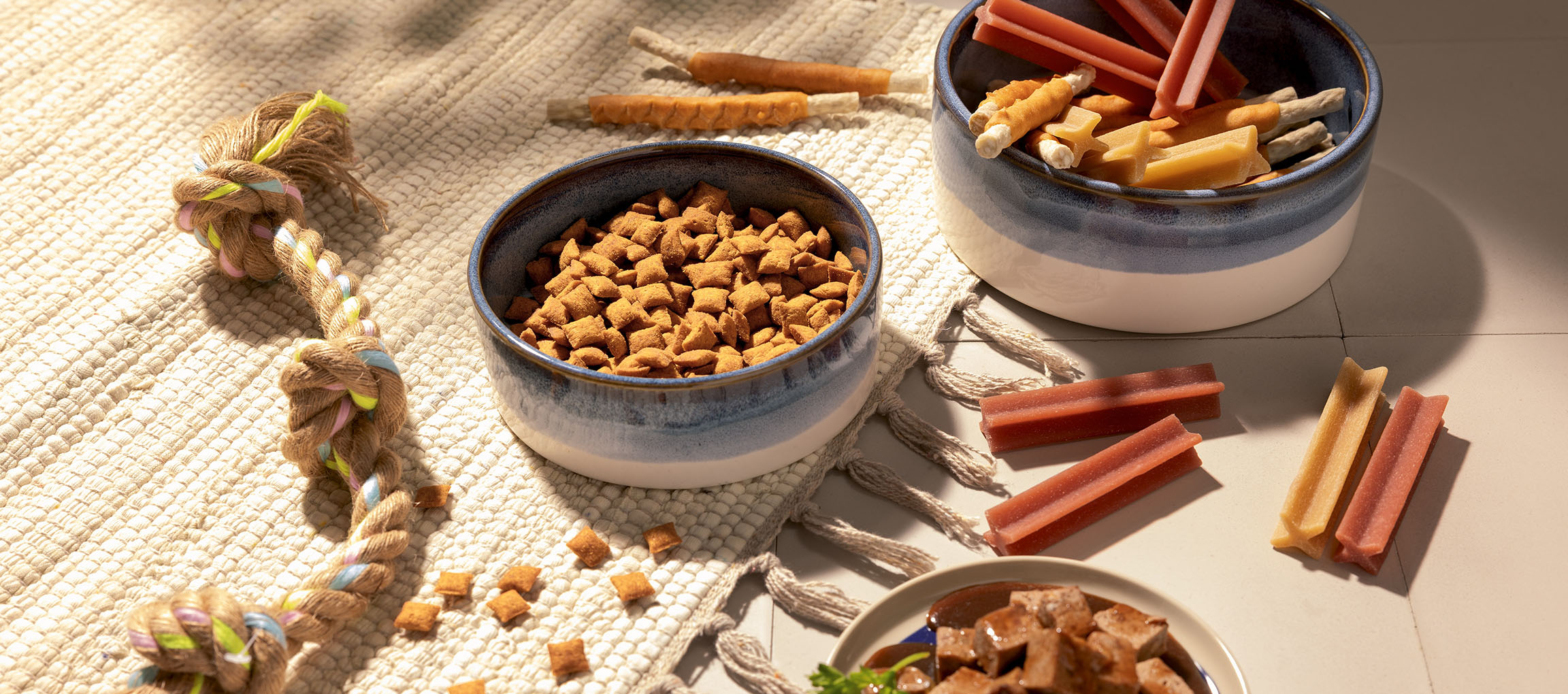 Cibo e snack assortiti per animali domestici, inclusi crocchette, bastoncini da masticare e un gioco di corda.